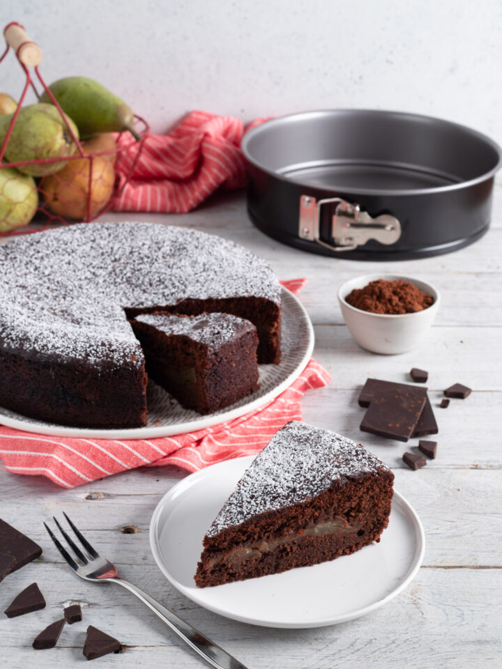 fetta di torta pere e cioccolato con tortiera sullo sfondo e frutta fresca