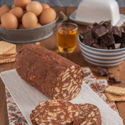 Salame di cioccolato su piatto rettangolare bianco, con due fette tagliate, bicchierino di liquore, cioccolato a cubetti e uova sullo sfondo