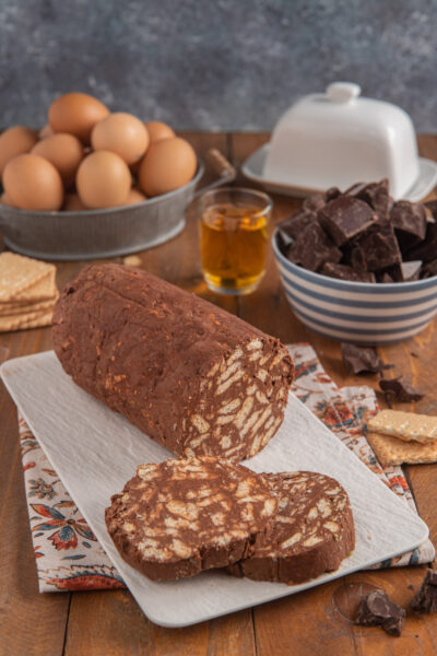 Salame di cioccolato su piatto rettangolare bianco, con due fette tagliate, bicchierino di liquore, cioccolato a cubetti e uova sullo sfondo