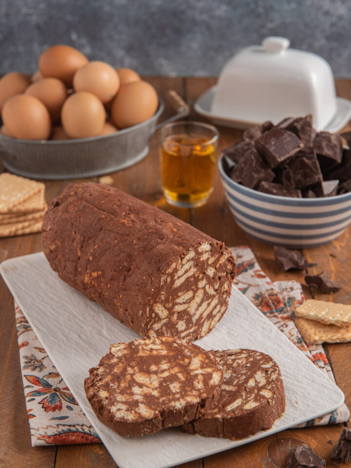Salame di cioccolato su piatto rettangolare bianco, con due fette tagliate, bicchierino di liquore, cioccolato a cubetti e uova sullo sfondo