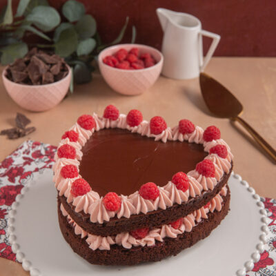 Torta cuore al cioccolato e lamponi, su piatto bianco decorato, con paletta da dolci, bricchetto e ciotolina di lamponi