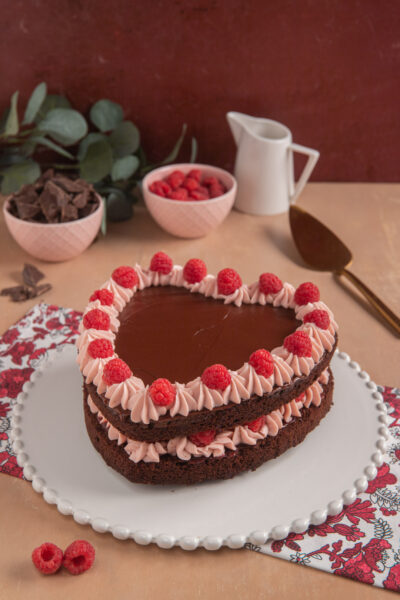 Torta cuore al cioccolato e lamponi, su piatto bianco decorato, con paletta da dolci, bricchetto e ciotolina di lamponi