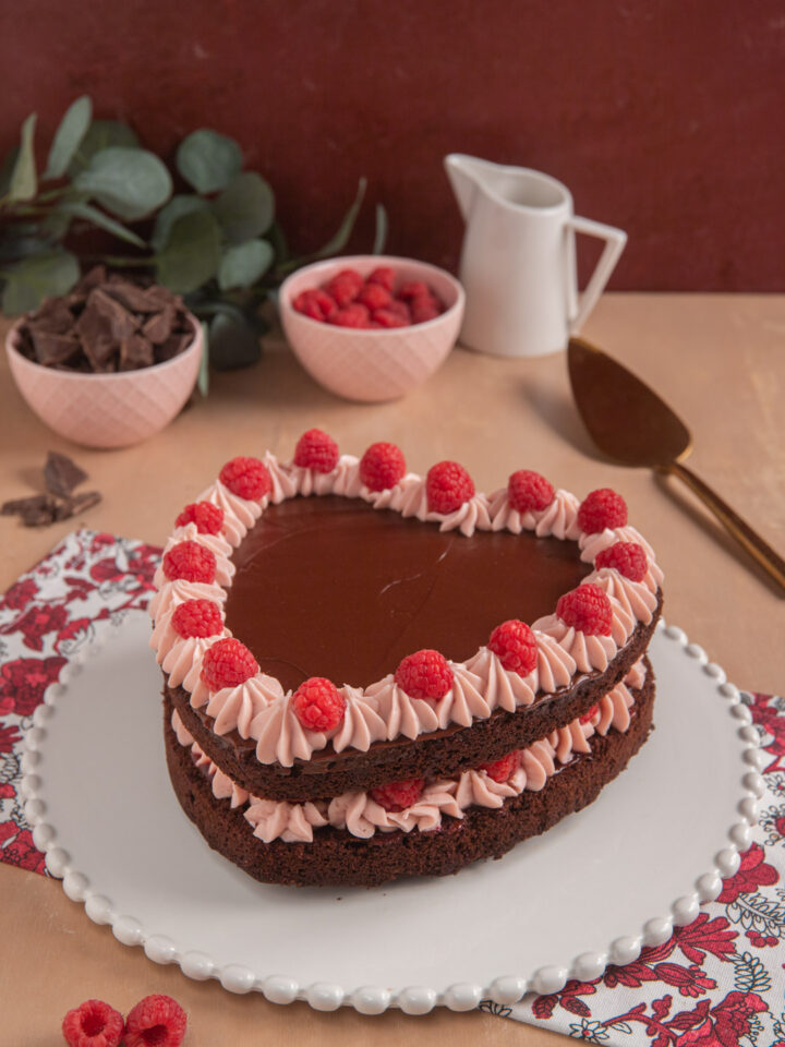 Torta cuore al cioccolato e lamponi, su piatto bianco decorato, con paletta da dolci, bricchetto e ciotolina di lamponi