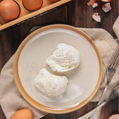 Uova in camicia impiattate, circondate da uova intere nel cartone, posate argentate e tovagliolo beige