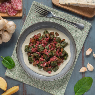 Gnocchetti agli spinaci con salame, funghi e gorgonzola su tovaglietta grigia, aglio e fette di salame