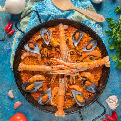 Paella de marisco nella tipica pentola di ferro, su sfondo azzurro, con cucchiaio di legno e ingredienti attorno