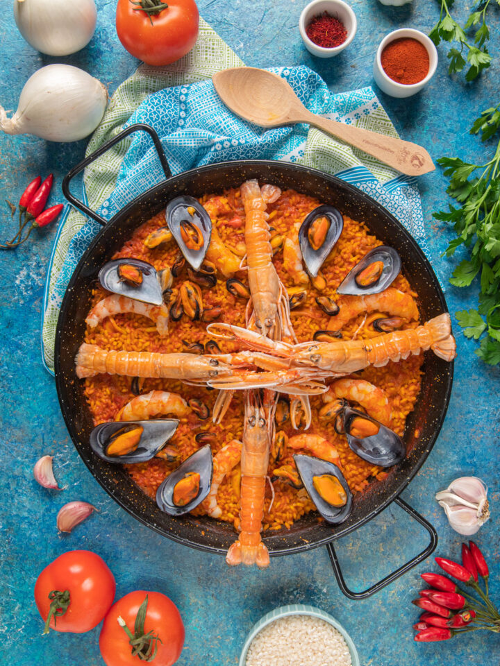 Paella de marisco nella tipica pentola di ferro, su sfondo azzurro, con cucchiaio di legno e ingredienti attorno