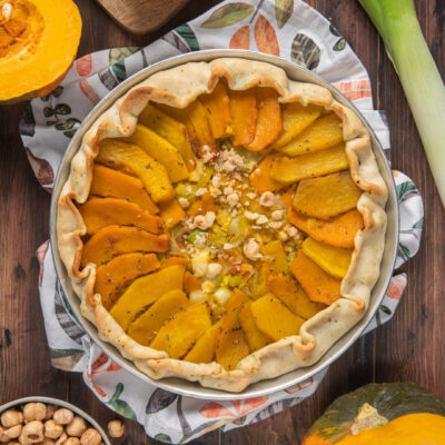 Torta salata zucca e nocciole nella tortiera con attorno zucca a metà e nocciole intere