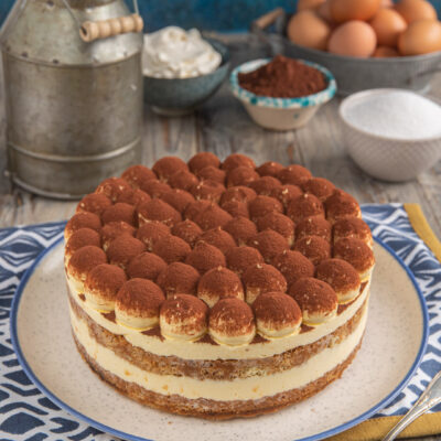 Torta tiramisù su tovaglietta bianca e blu con ciotoline di cacao e zucchero e uova