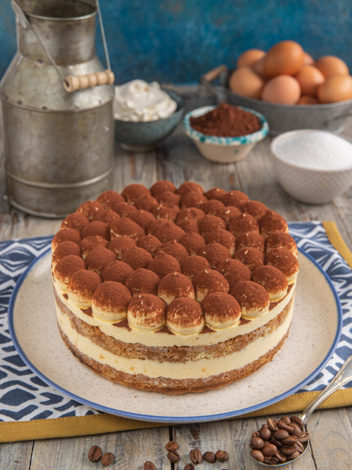 Torta tiramisù su tovaglietta bianca e blu con ciotoline di cacao e zucchero e uova