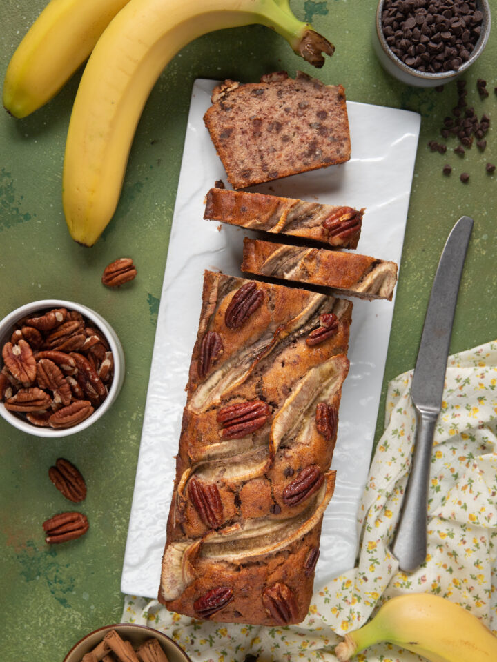 banana bread tagliato a fette su tagliere con intorno banane, cioccolato e noci pecan