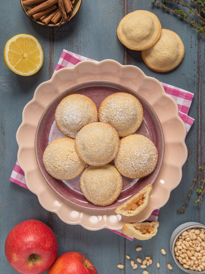 Biscotti alle mele su piatto rosa con limone, cannella, mele e pinoli, su tovagliolo a quadretti bianchi e rossi