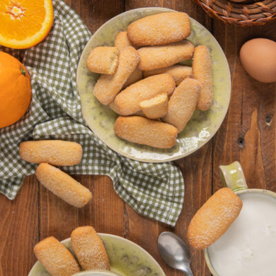 Biscotti da inzuppo in piattini verdi, con canovaccio a quadretti, tazza di latte e arancia a metà