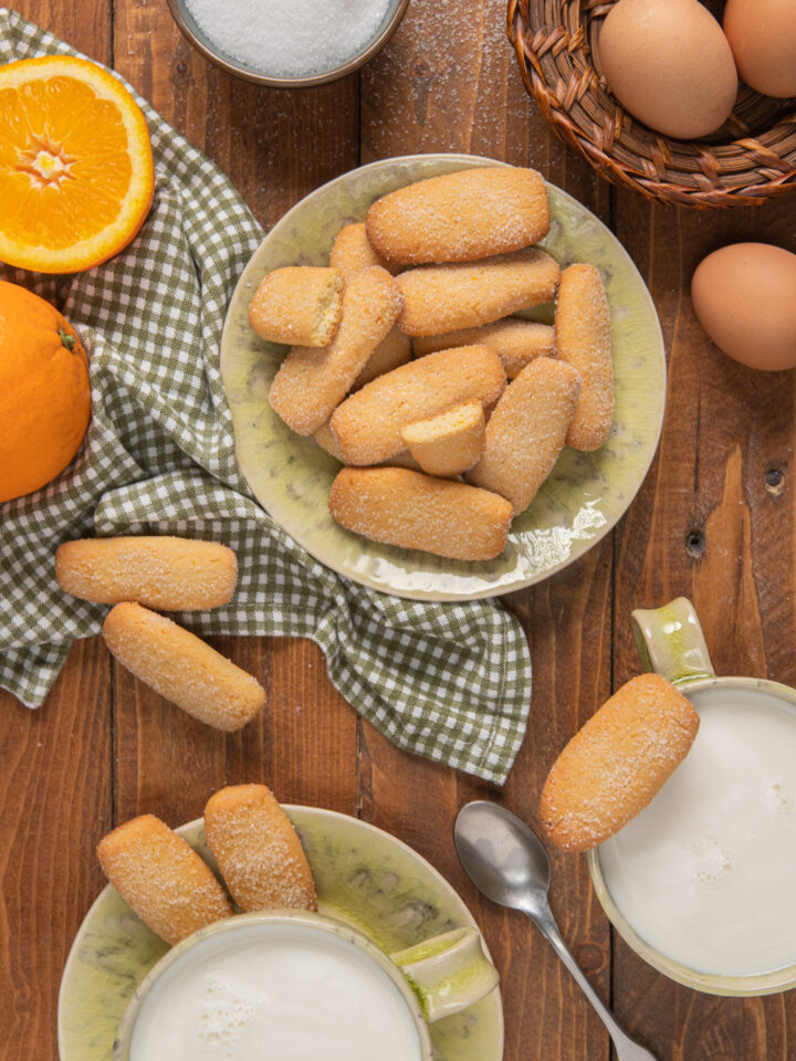 Biscotti da inzuppo in piattini verdi, con canovaccio a quadretti, tazza di latte e arancia a metà