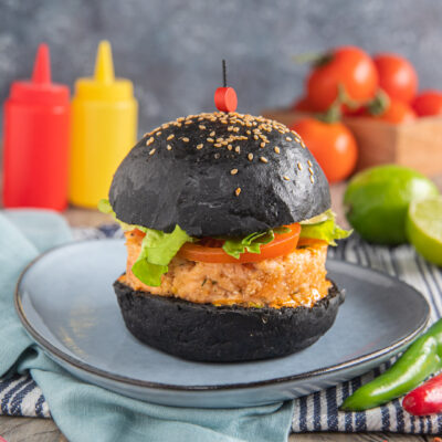 Burger di salmone con bun al carbone su piatto grigio, con biberon rosso e giallo sullo sfondo, lime e pomodori