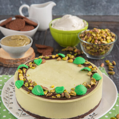Cheesecake al pistacchio su piatto decorato con foglie, ciotoline sullo sfondo e tovaglietta verde a pallini bianchi