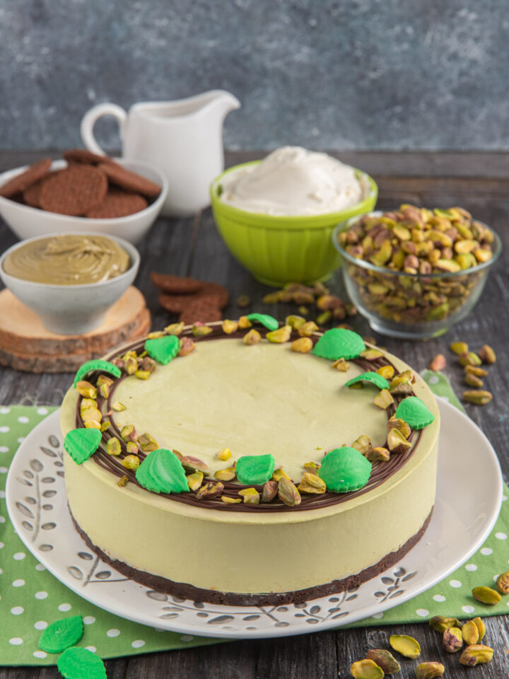 Cheesecake al pistacchio su piatto decorato con foglie, ciotoline sullo sfondo e tovaglietta verde a pallini bianchi