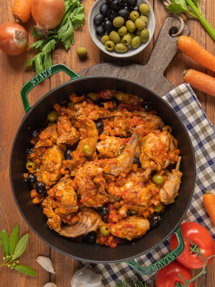 Coniglio alla cacciatora in pentola, su tagliere di legno, canovaccio a quadretti, carote, pomodori, ciotolina di olive verdi ed erbe aromatiche