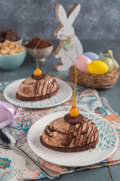 Gelato di Pasqua con sablè al cacao su due piattini, con tovagliette colorate, cestino di uova colorate e coniglietto sullo sfondo