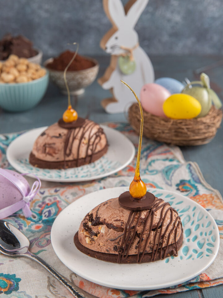 Gelato di Pasqua con sablè al cacao su due piattini, con tovagliette colorate, cestino di uova colorate e coniglietto sullo sfondo