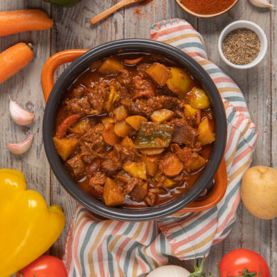 Gulash ungherese (Gulyás) in pentola con manici arancioni, tovagliolo a righe colorate , peperone giallo, carota, pomodori, patata e cipolla