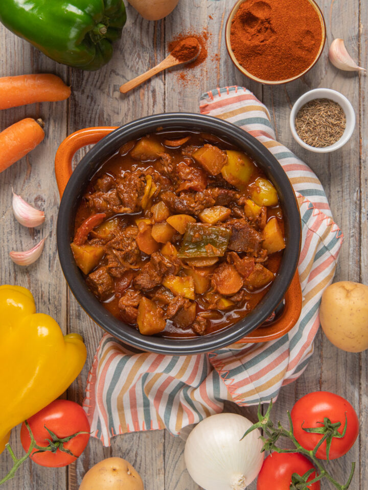 Gulash ungherese (Gulyás) in pentola con manici arancioni, tovagliolo a righe colorate , peperone giallo, carota, pomodori, patata e cipolla