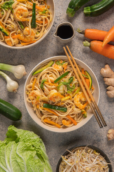 Noodles con verdure e gamberi in due ciotole con verdure crude, bacchette e bricchetto con salsa di soia