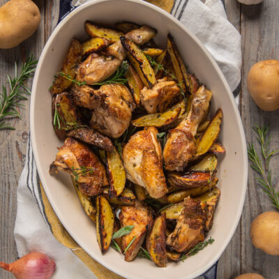 Pollo al forno con patate in teglia ovale bianca con patate cotte a spicchi e patate crude intere, rosmarino e scalogni