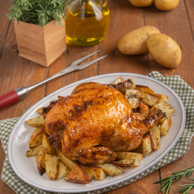 Pollo arrosto con patate cotte a spicchi su piatto da portata ovale e tovagliolo a quadretti, più forchettone e patate crude