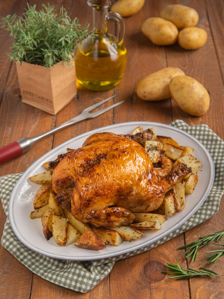 Pollo arrosto con patate cotte a spicchi su piatto da portata ovale e tovagliolo a quadretti, più forchettone e patate crude