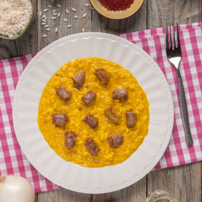 Risotto alla monzese in piatto bianco su runner a quadri bianchi e rossi, con ciotoline di pistilli di zafferano e riso, e cipolle intere