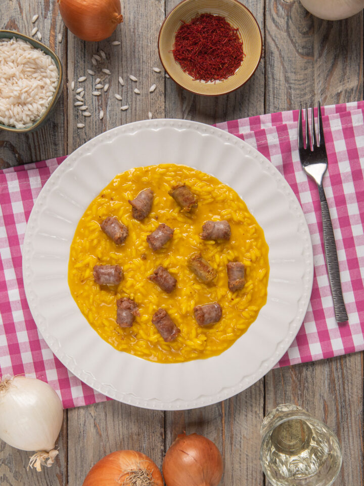 Risotto alla monzese in piatto bianco su runner a quadri bianchi e rossi, con ciotoline di pistilli di zafferano e riso, e cipolle intere