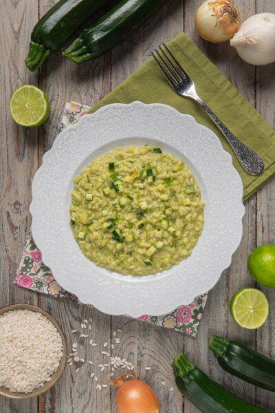 Foto risotto alle zucchine su piatto bianco, tovaglietta verde e contorno di zucchine crude, cipolle, lime a metà e ciotolina di riso