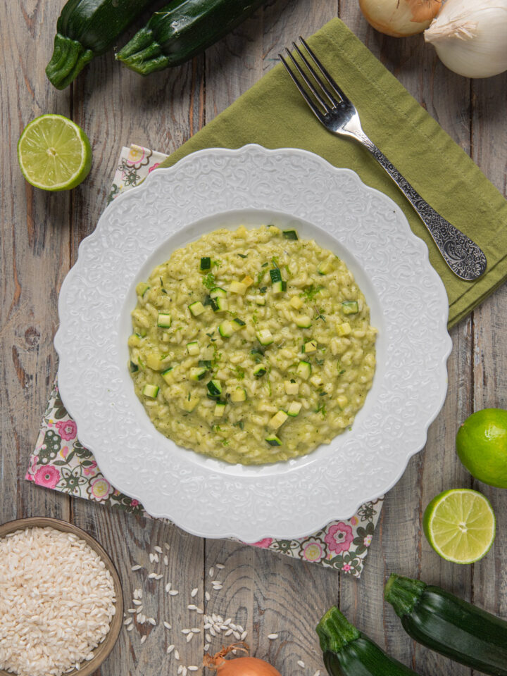 Foto risotto alle zucchine su piatto bianco, tovaglietta verde e contorno di zucchine crude, cipolle, lime a metà e ciotolina di riso