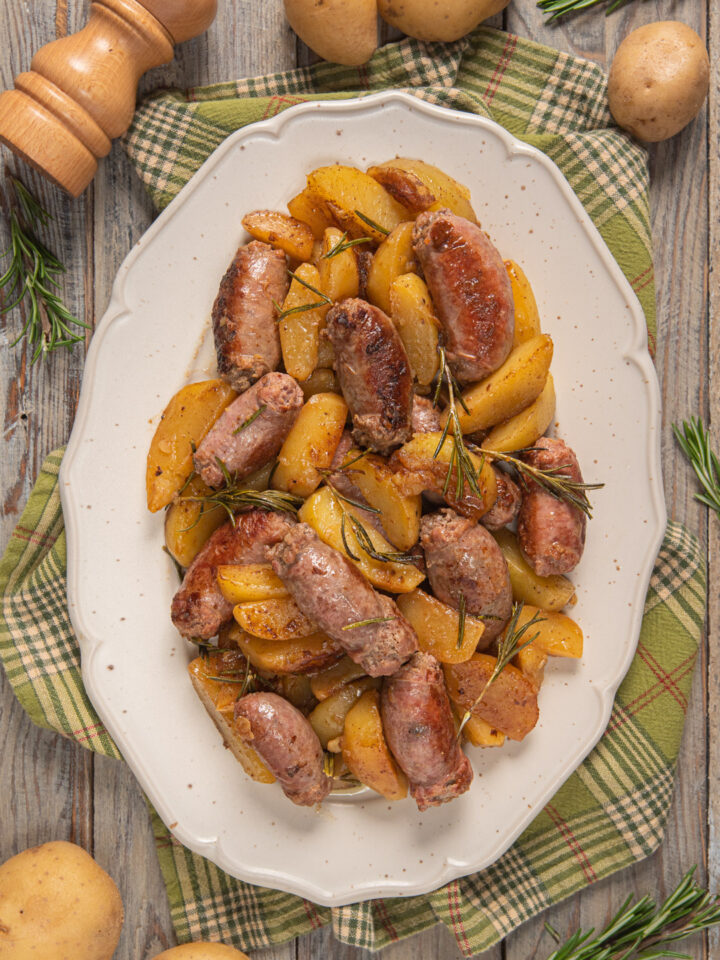 Salsiccia e patate in padella, impiattate in piatto ovale bianco con tovaglietta verde a righe perpendicolari