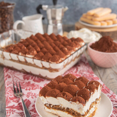 Tiramisù senza uova con fetta tagliata, tovaglietta rosa, caffettiera sullo sfondo e ciotolina di cacao