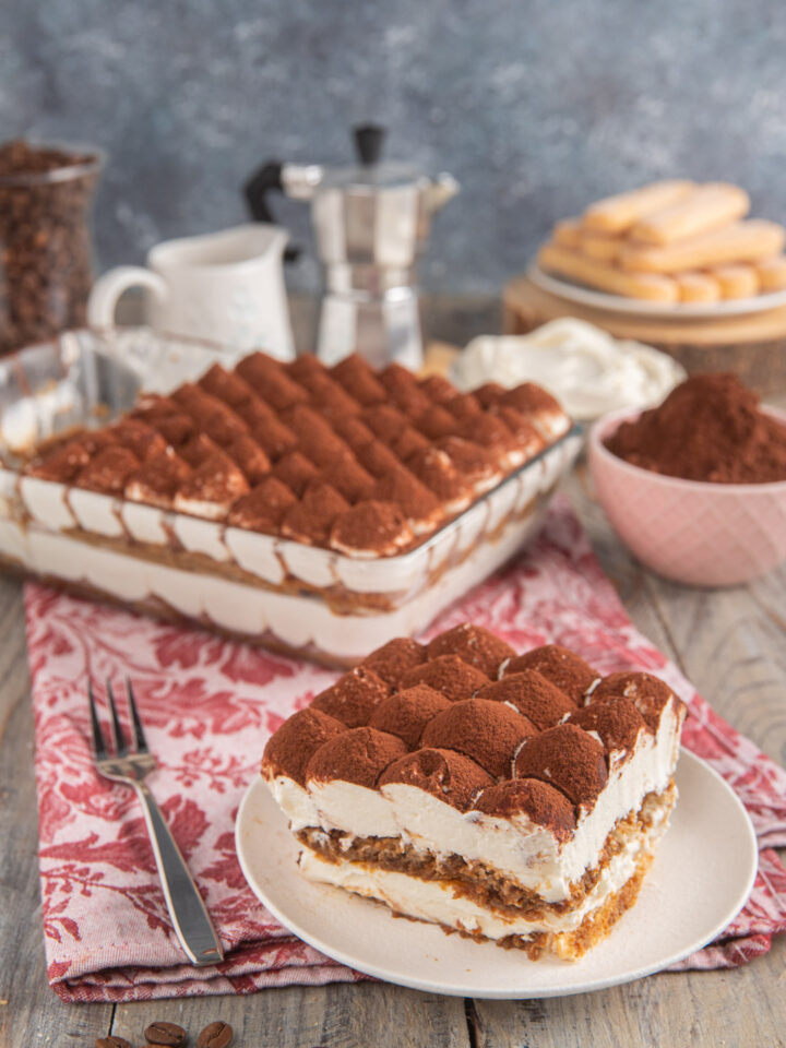 Tiramisù senza uova con fetta tagliata, tovaglietta rosa, caffettiera sullo sfondo e ciotolina di cacao