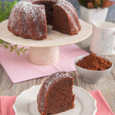 Torta al cacao senza uova su alzatina bianca, con fetta tagliata su piatto bianco, tovaglioli rosa e cotolina di cacao.