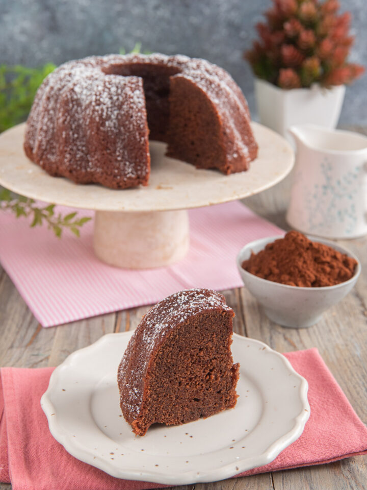 Torta al cacao senza uova su alzatina bianca, con fetta tagliata su piatto bianco, tovaglioli rosa e cotolina di cacao.