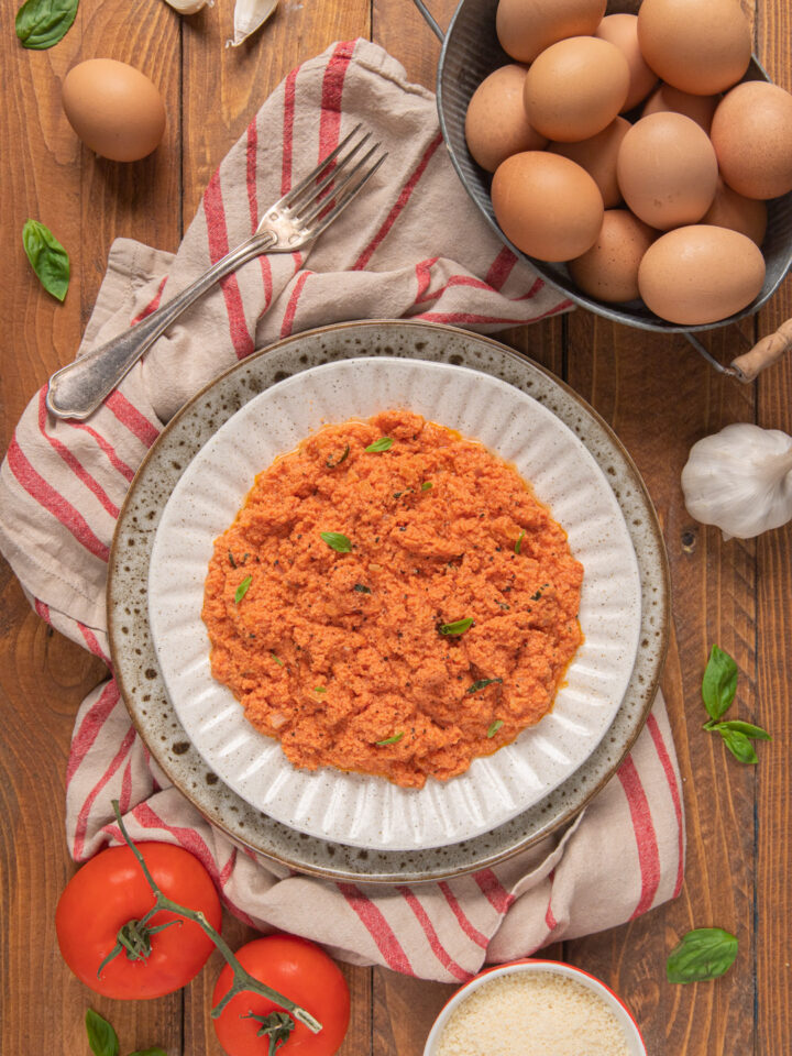 Uova strapazzate al pomodoro su canovaccio bianco a righe rosse, cestino di uova, pomodoro, aglio e foglioline di basilico