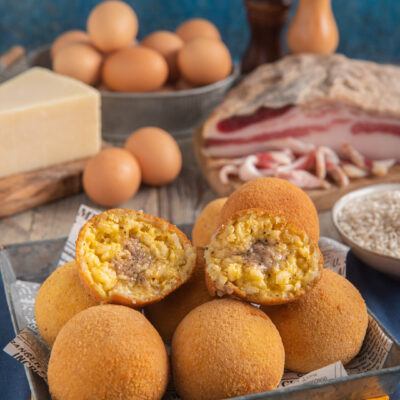 Arancini alla carbonara interi e divisi a metà con pezzo intero di pecorino sullo sfondo, uova e guanciale