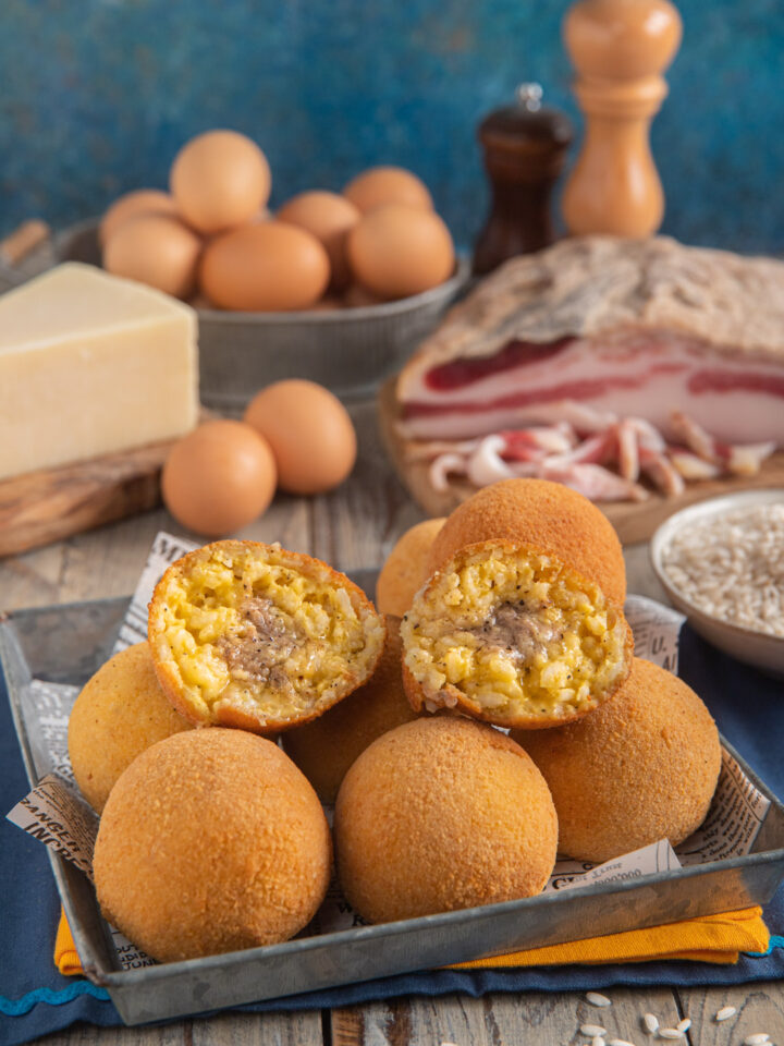 Arancini alla carbonara interi e divisi a metà con pezzo intero di pecorino sullo sfondo, uova e guanciale