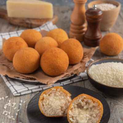 Arancini cacio e pepe impiattati su carta-paglia con tovagliolo a scacchi, con un arancino diviso a metà in primo piano, ciotolina di riso e pezzo di pecorino intero sullo sfondo