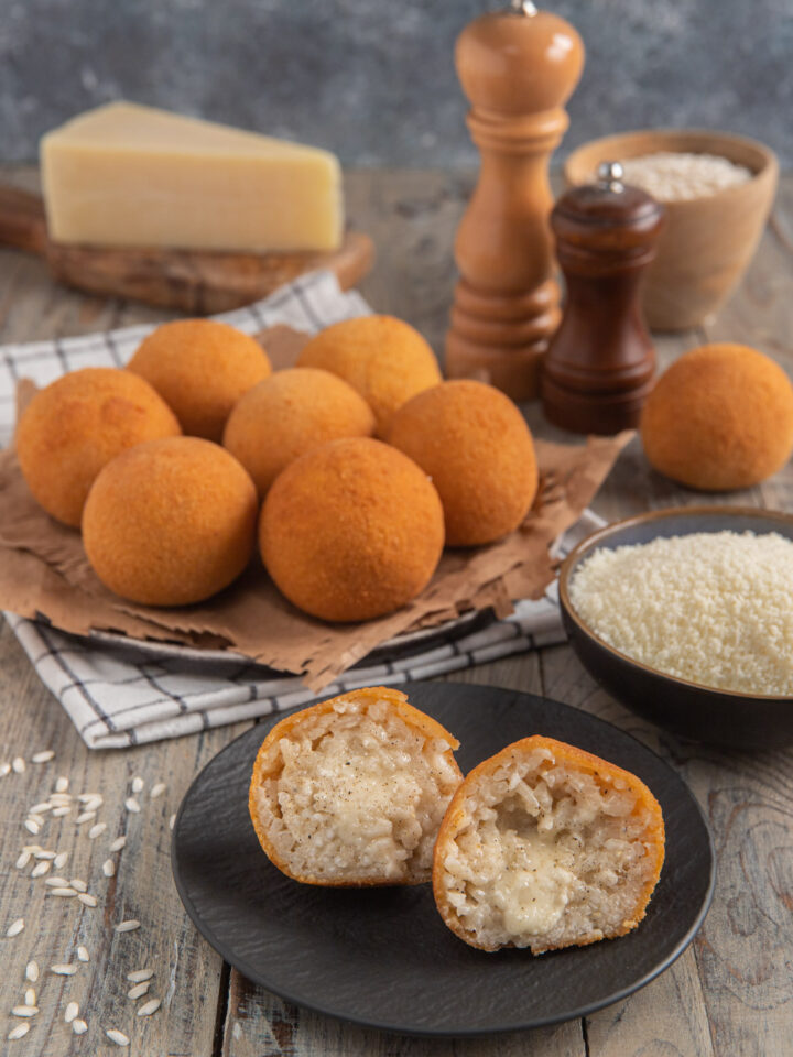 Arancini cacio e pepe impiattati su carta-paglia con tovagliolo a scacchi, con un arancino diviso a metà in primo piano, ciotolina di riso e pezzo di pecorino intero sullo sfondo