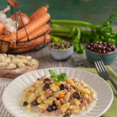 Cavatelli al ragù di cortile impiattati in piatto bianco con bordo decorato, con sullo sfondo carote, capperi, olive e sedano