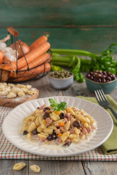 Cavatelli al ragù di cortile impiattati in piatto bianco con bordo decorato, con sullo sfondo carote, capperi, olive e sedano