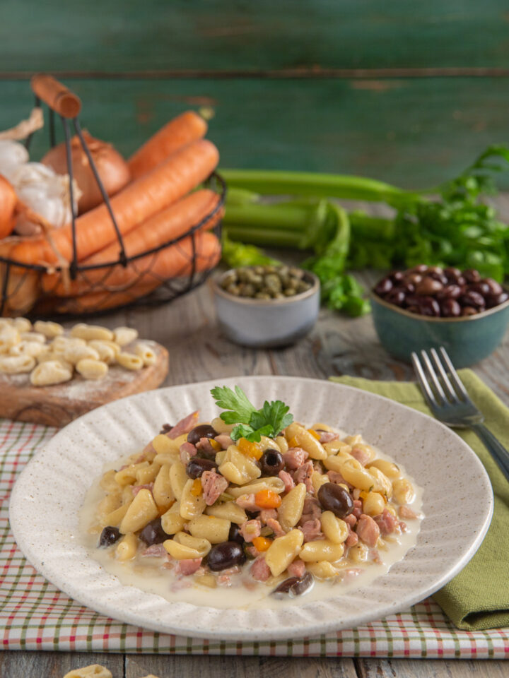 Cavatelli al ragù di cortile impiattati in piatto bianco con bordo decorato, con sullo sfondo carote, capperi, olive e sedano