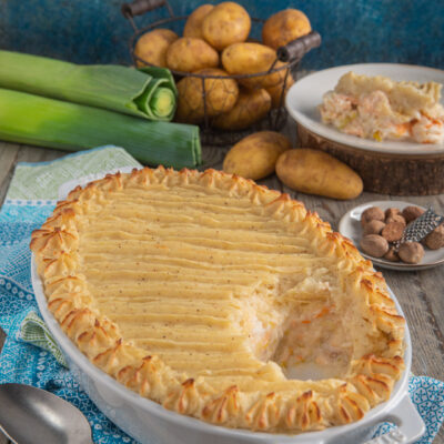 Fish pie in teglia ovale su canovaccio azzurro, con porri e patate sullo sfondo e piattino con porzione tagliata