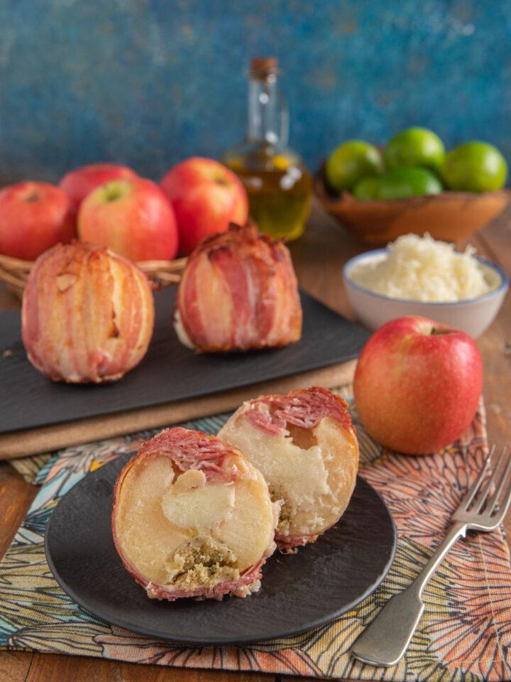 Mele con bacon e pecorino con mela in primo piano divisa a metà su piattino nero e forchettina a destra, più due mele intere farcite sullo sfondo