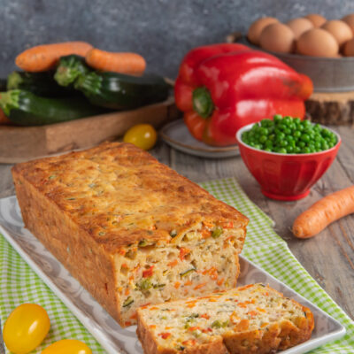 Torta salata con yogurt e verdure su piatto rettangolare, con una fetta tagliata, pomodorini gialli, ciotolina di piselli e peperone rosso sullo sfondo